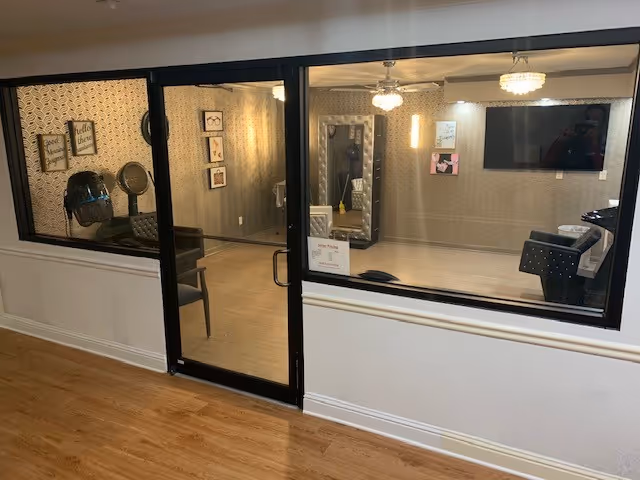 Interior view through glass windows and a door into a room with wooden flooring, a wall-mounted TV, framed pictures on the walls, a salon hair dryer chair, and modern lighting fixtures on the ceiling.