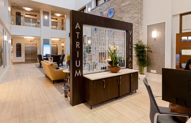 Interior view of a senior living facility lobby with a modern wooden divider featuring the words 'ATRIUM VILLAGE'. The space includes comfortable seating areas with chairs and sofas, light-colored tiled flooring, a stone accent wall with decorative plates, potted plants, and an elevator in the background.