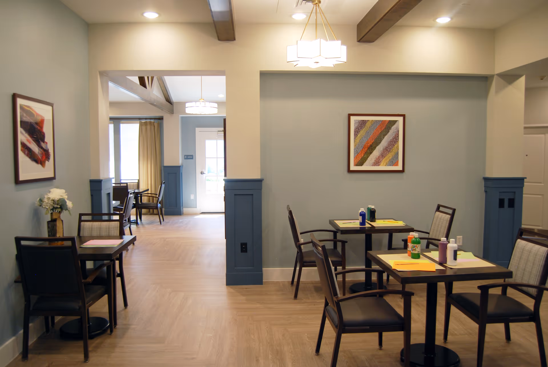 Interior view of a senior living facility dining area with several small tables and chairs. The walls are painted light blue with darker blue wainscoting. There are framed abstract artworks on the walls, ceiling lights, and a vase with white flowers on one table. The room has a wooden floor and an open doorway leading to another similar dining space with more tables and chairs.