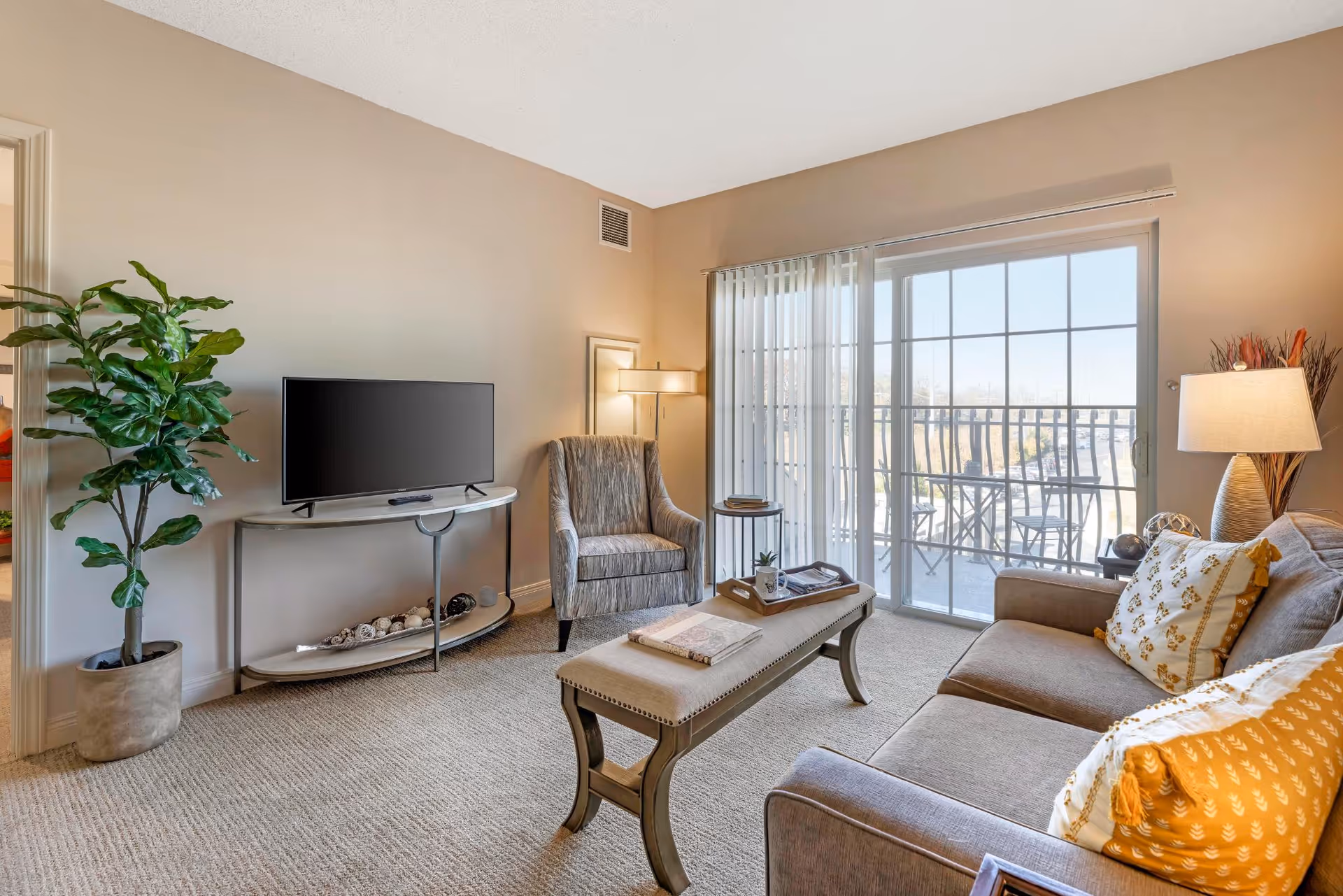 A cozy living room with a gray sofa adorned with yellow and white patterned pillows, a cushioned bench-style coffee table, a gray armchair, a TV on a curved stand, a tall potted plant, and a sliding glass door leading to a balcony with outdoor seating.