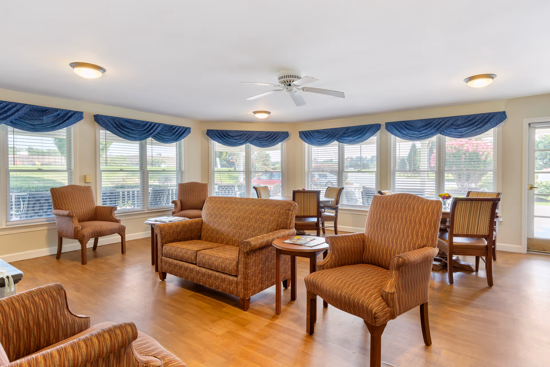 A bright and spacious common area with multiple upholstered chairs and a loveseat arranged around small wooden tables. Large windows with blue valances let in natural light and offer views of greenery outside. The room has a ceiling fan and wood flooring.