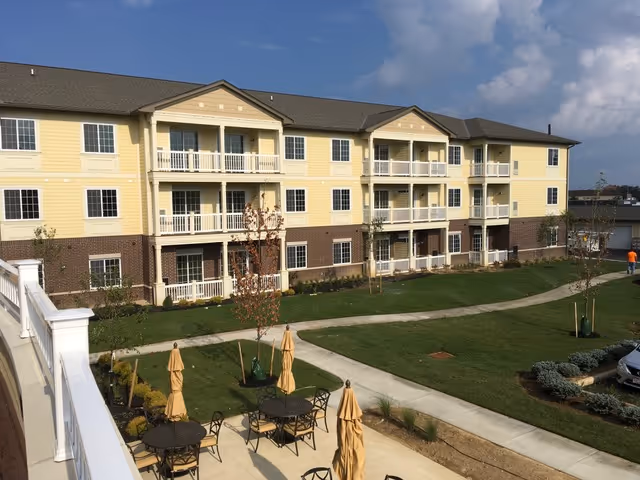 Exterior view of a three-story senior living facility building with yellow siding and brick accents. The building has balconies on the second and third floors. In the foreground, there is a landscaped lawn with a paved walkway, several patio tables with closed umbrellas, and some small trees and shrubs.
