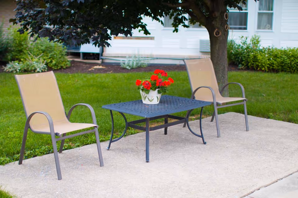 Two beige mesh chairs with metal frames are placed on a concrete patio around a black metal table with a lattice design. A white pot with red flowers sits on the table. Behind the patio is a green lawn, a tree providing shade, and a white building with windows and bushes.