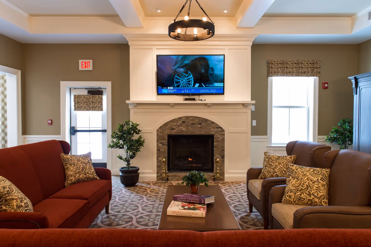 A cozy living room with a red sofa and two brown armchairs, each with patterned cushions. A wooden coffee table with books and a small plant sits in the center. A fireplace with a brick surround is centered on the far wall, above which a flat-screen TV is mounted. Two windows with patterned valances flank the fireplace, and there are potted plants on either side. A chandelier hangs from the ceiling, and an exit door is visible on the left side of the room.