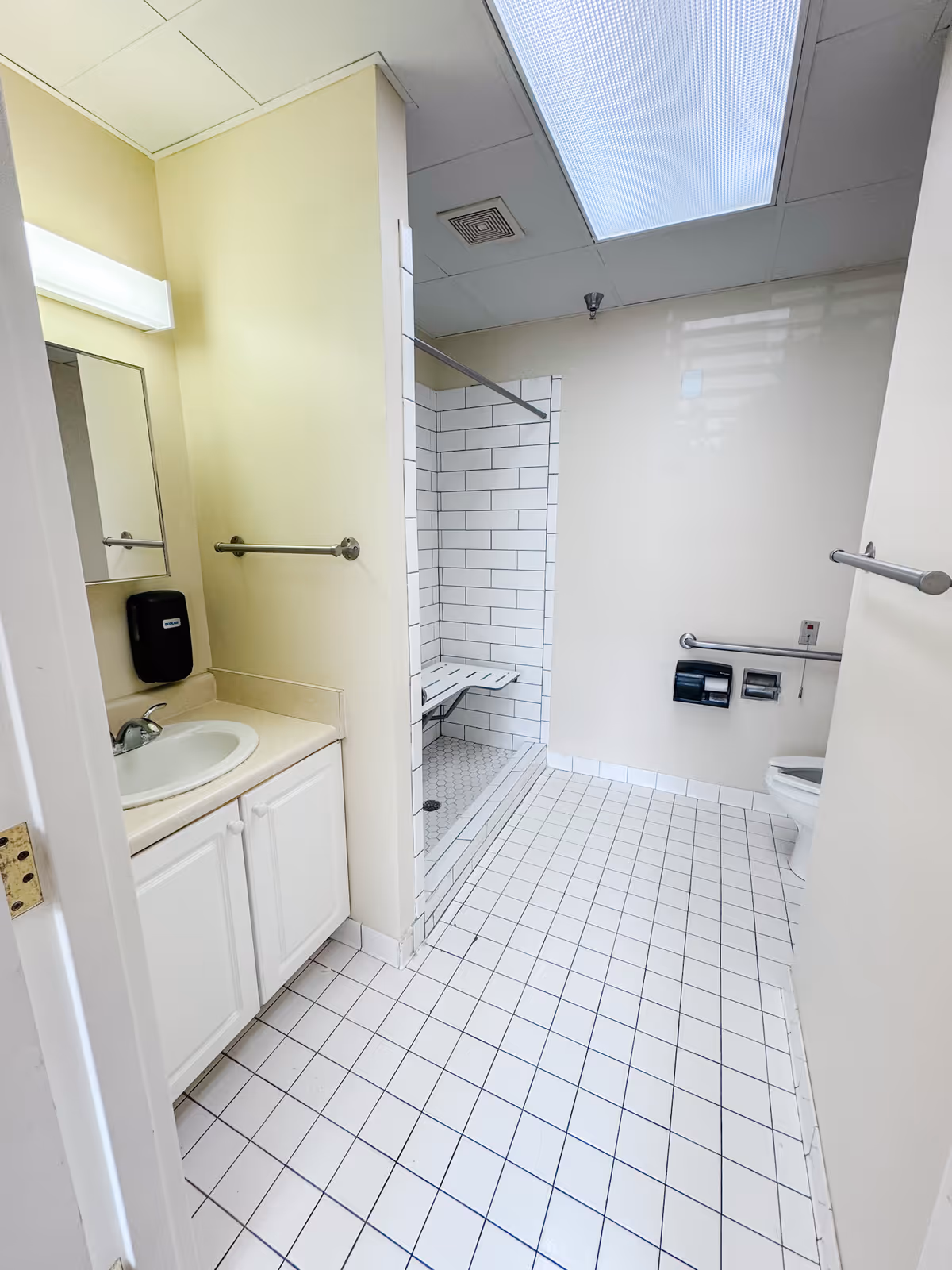 A clean, accessible bathroom with white tiled floor and walls. The room features a sink with a countertop and cabinet, a wall-mounted soap dispenser, a mirror, a shower area with a foldable seat and grab bars, and a toilet with grab bars and dual toilet paper holders. The ceiling has a large fluorescent light panel.