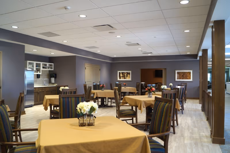Spacious dining room with multiple tables draped in yellow tablecloths, wooden chairs, floral centerpieces, and a small kitchen area along the back wall.