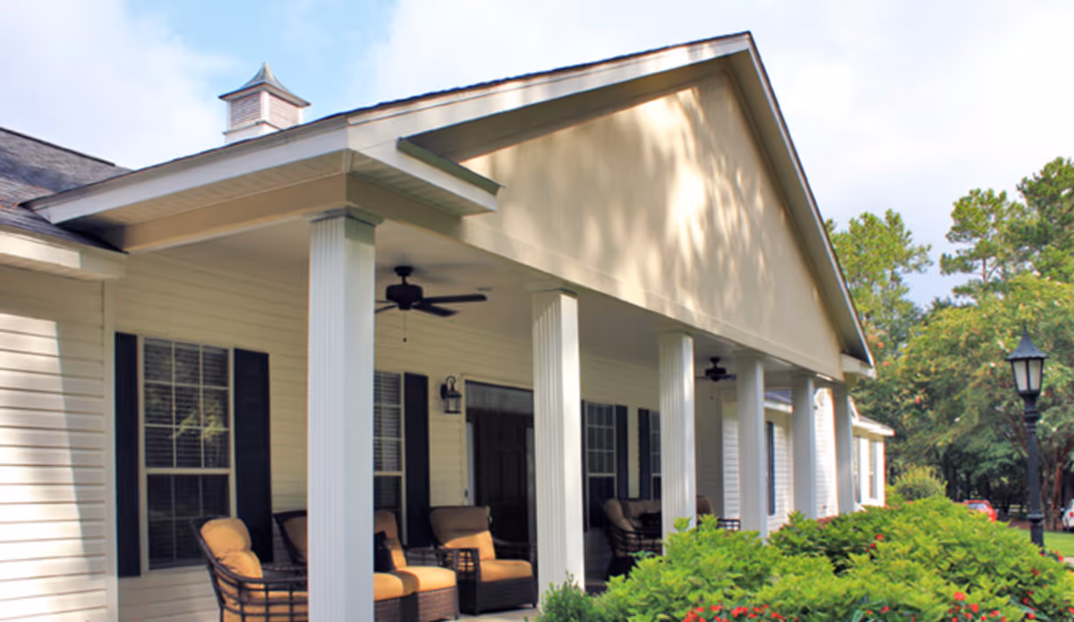 Exterior view of a single-story building with a covered porch supported by white columns. The porch has cushioned chairs and ceiling fans. There are windows with black shutters and a lamp post near green bushes and trees.