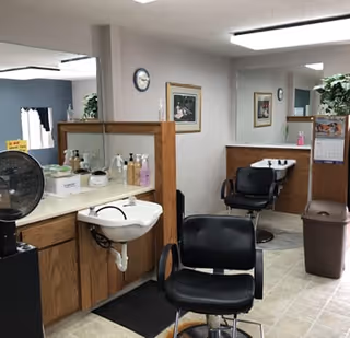 Interior of a hair salon area with two black salon chairs in front of sinks and wooden cabinets. There are mirrors on the walls, a clock, framed pictures, and various hair care products on the counters. A trash bin and a calendar are also visible.