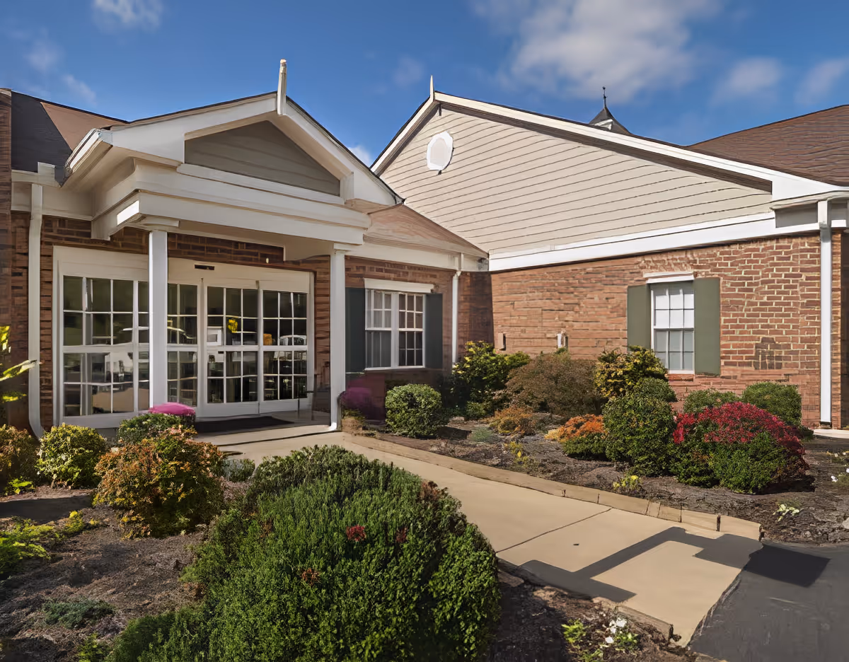 Exterior view of Charter Senior Living of Paris showing the entrance with glass double doors, brick walls, and a small garden with shrubs and bushes along the walkway under a blue sky with some clouds.