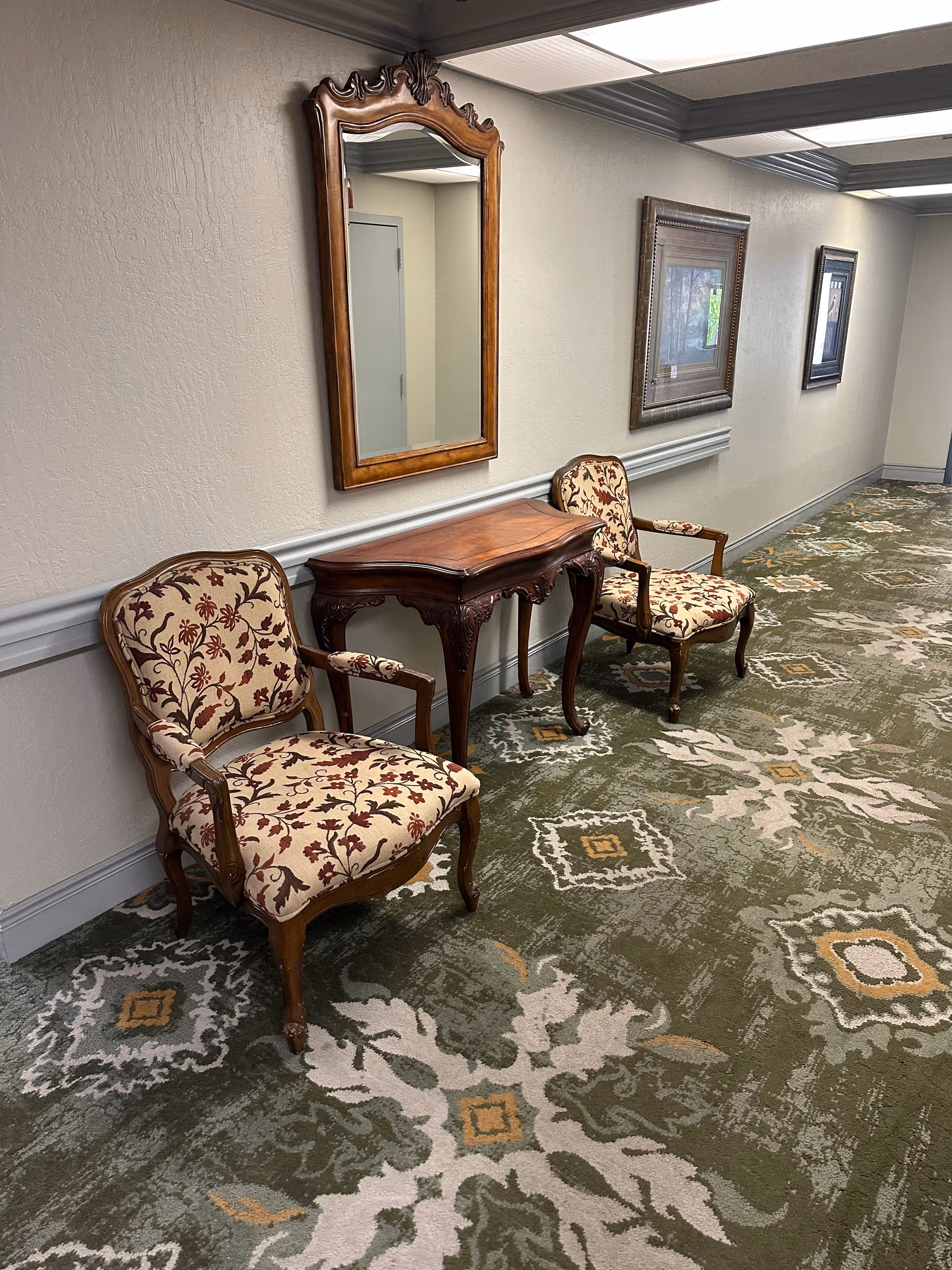 A hallway with two upholstered armchairs featuring floral patterns, a wooden console table with carved legs, and a large ornate mirror hanging on the wall above the table. The hallway has patterned green carpet and framed artwork on the walls.