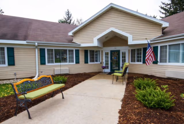 Exterior view of a single-story beige building with green shutters and a brown roof. There is a concrete walkway leading to a glass door entrance with a small overhang. Two green chairs and a bench with a green cushion are placed near the entrance. An American flag is mounted on the right side of the entrance. Small bushes and mulch line the walkway.