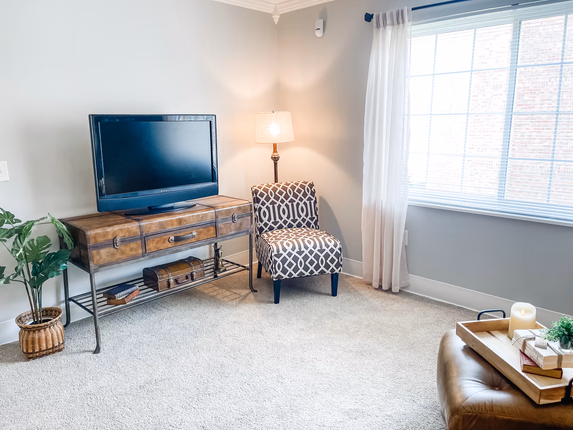 A cozy living room corner with a patterned armchair next to a lit floor lamp, a wooden TV stand with a flat-screen television, a potted plant on the floor, and a large window with sheer curtains letting in natural light. A leather ottoman with a wooden tray holding a candle, books, and a small plant is partially visible.