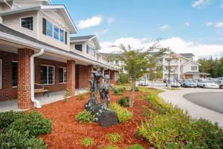 Exterior view of Dogwood Terrace by Barclay House showing a brick building with white trim, a landscaped garden area with a bronze statue, small trees, and shrubs, and a curved driveway with parked cars under a partly cloudy sky.