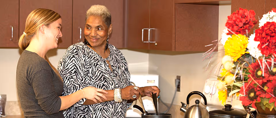 Two women smiling and cooking together in a kitchen. One woman is younger with light skin and blonde hair tied back, and the other is older with short gray hair wearing a zebra-patterned top. They are standing near a stove with pots and a kettle, and there is a colorful flower arrangement on the right side of the image.