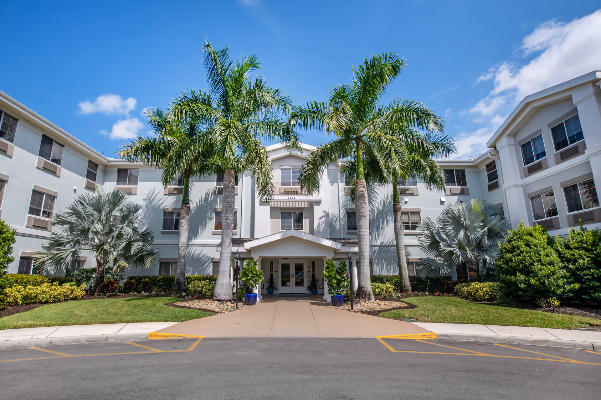 Front entrance of a three-story white senior living building framed by tall palm trees and landscaping under a blue sky.
