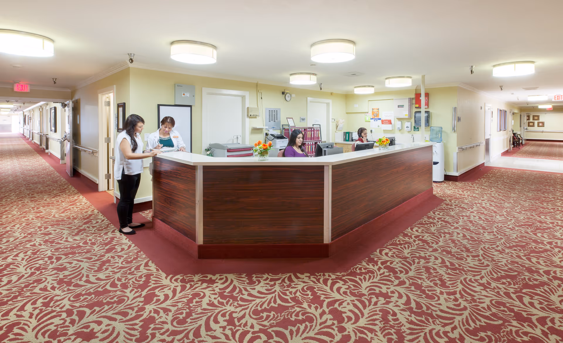 Reception area in a healthcare facility with a large wooden front desk. Two staff members are seated behind the desk working on computers, while two other people stand in front of the desk engaged in conversation. The area has patterned red carpet, cream-colored walls, and bright ceiling lights. Hallways extend on both sides of the reception desk.