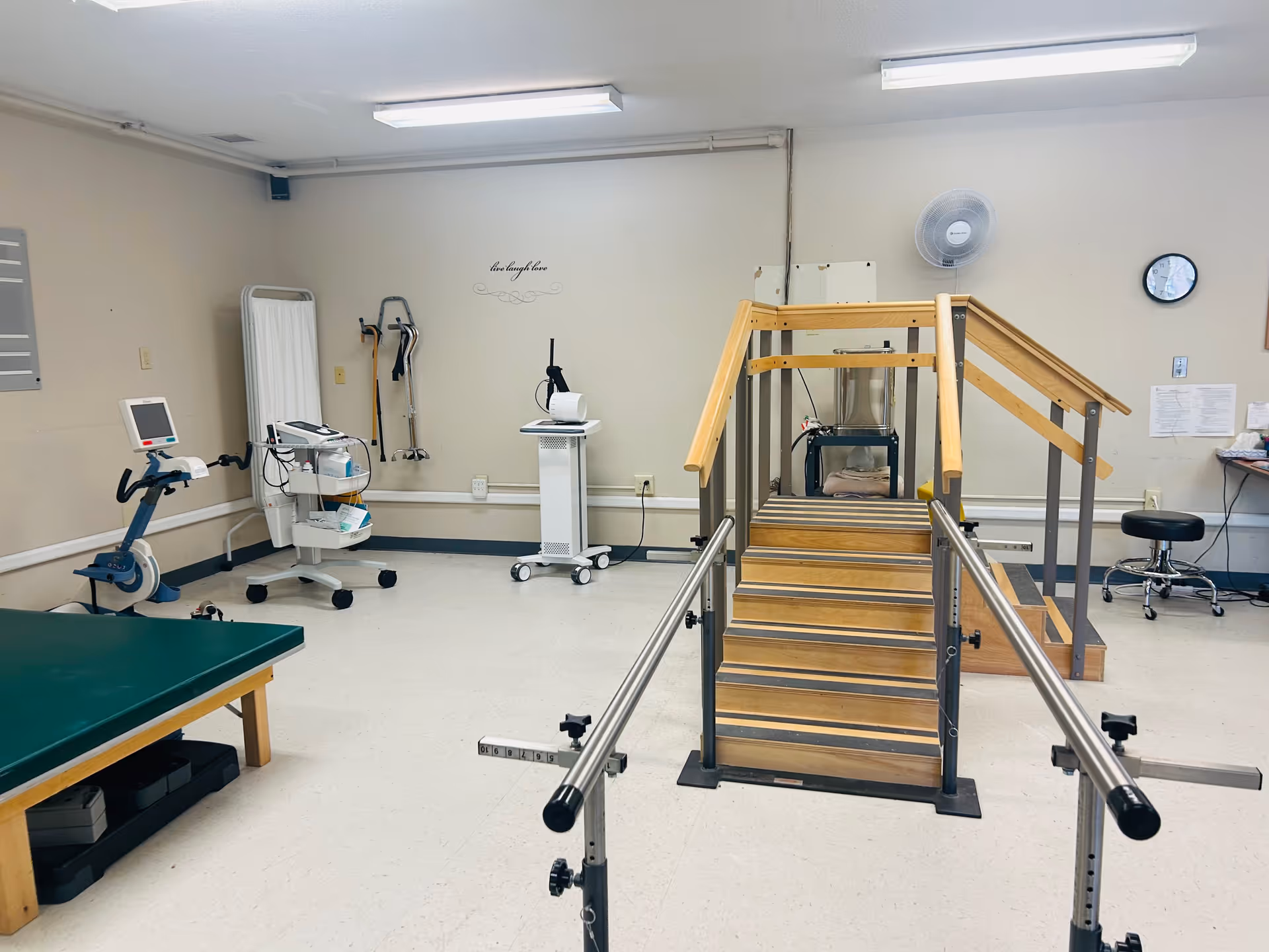 A rehabilitation therapy room with parallel bars and a wooden staircase for physical therapy exercises. The room also contains medical equipment, a green therapy table, a stool, and a wall clock. The walls are beige, and there is a motivational decal that reads 'live laugh love'.