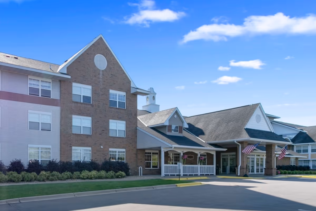 Exterior view of St. Andrew’s Village senior living facility showing a multi-story building with brick and siding facade, a covered entrance with white railings, American flags, and a clear blue sky.