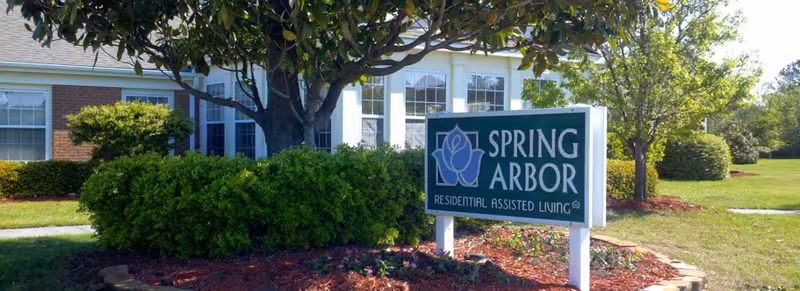 Outdoor view of the Spring Arbor Residential Assisted Living facility sign surrounded by green bushes and trees, with part of the building visible in the background under a clear sky.