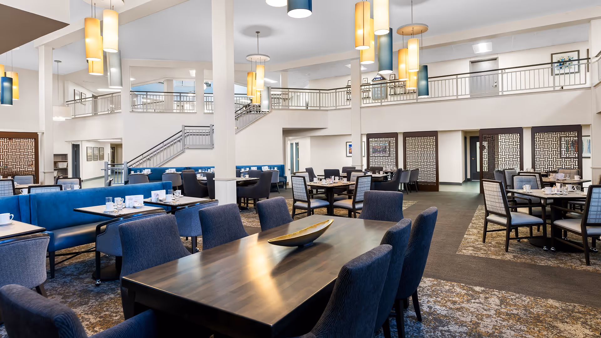 Spacious dining room with multiple tables and chairs arranged neatly, featuring blue cushioned seating and modern hanging pendant lights. The room has a high ceiling with a second-floor balcony and staircase visible in the background.