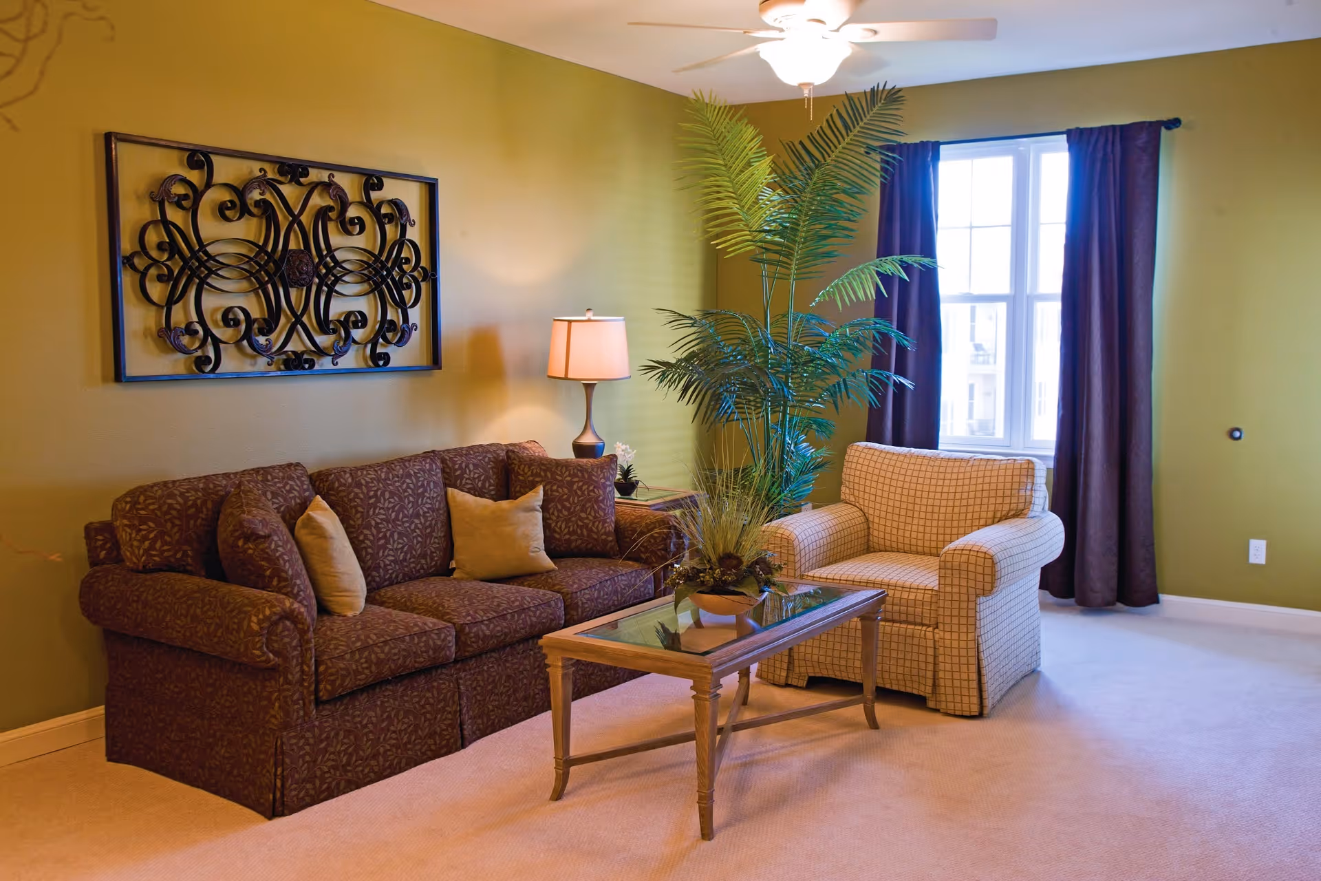 A cozy living room with a patterned brown sofa and a beige checkered armchair. A glass-top wooden coffee table with a potted plant sits in front of the seating. There is a tall green plant next to the armchair, a table lamp on a side table, and a decorative wrought iron wall art hanging on the olive green wall. A window with dark curtains lets in natural light.