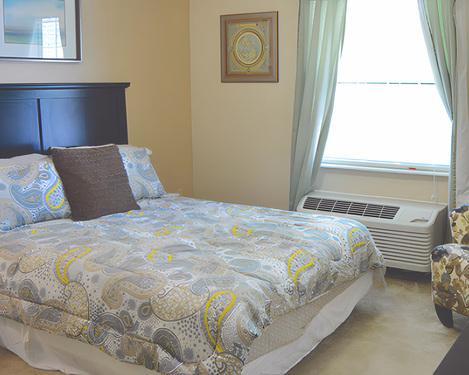 A furnished bedroom with a patterned bed, dark headboard, window with curtains and a wall air-conditioning unit.