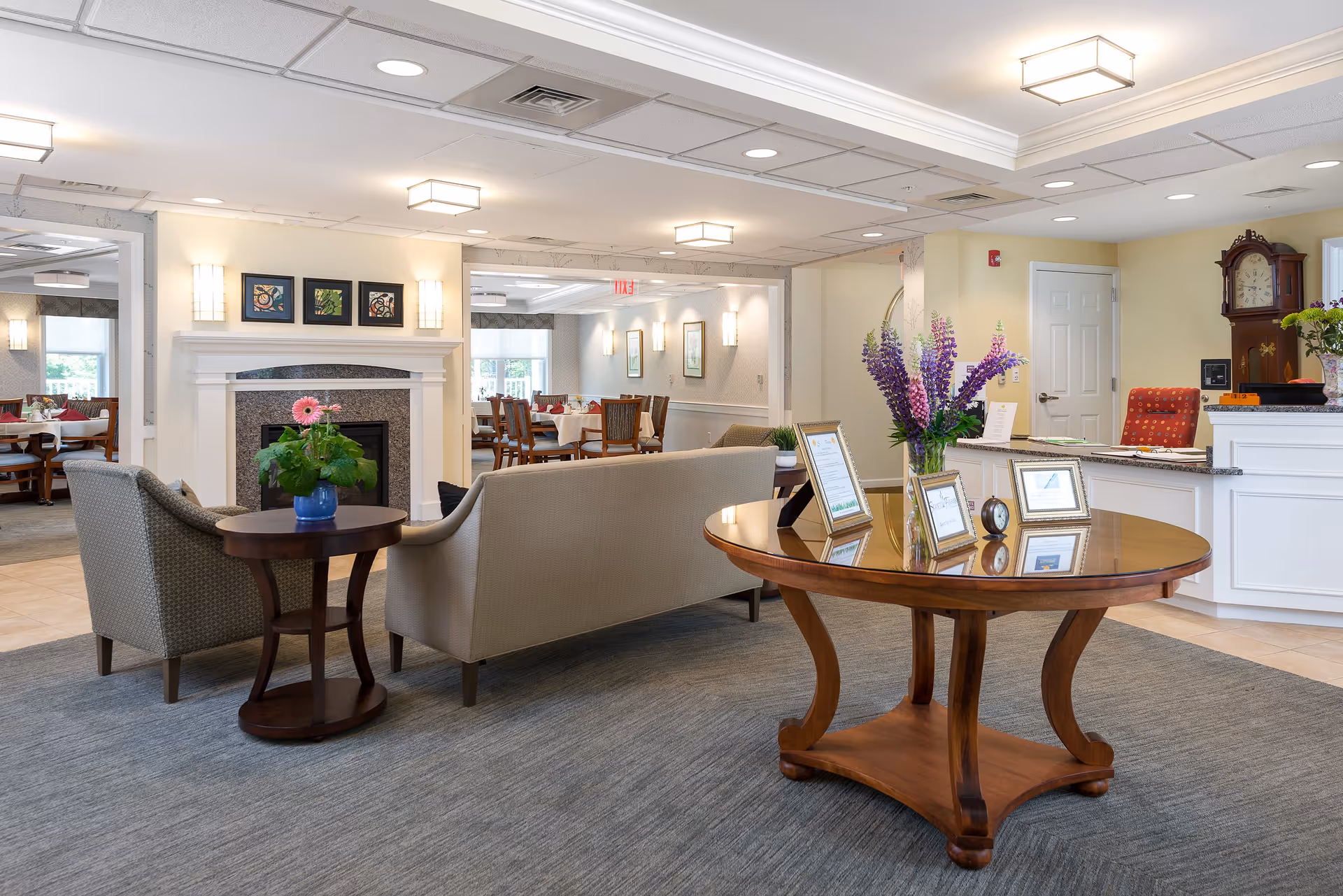 A bright and welcoming senior living facility common area with a beige sofa and armchair around a small round wooden table with a potted plant. In the foreground, a larger round wooden table holds a vase of purple flowers and several framed documents. The background shows a fireplace with three framed artworks above it and a dining area with tables and chairs. The reception desk with a red chair and a grandfather clock is visible to the right.