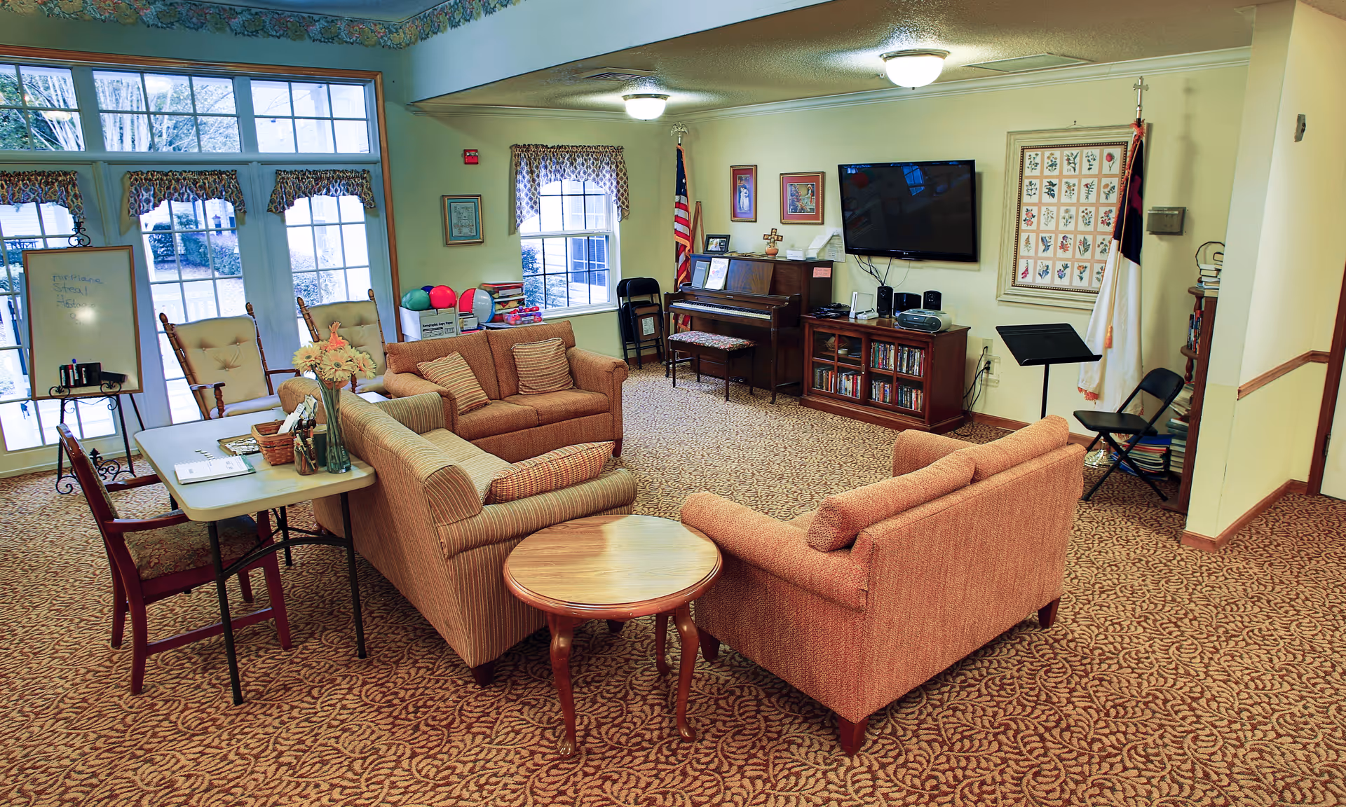 A cozy senior living lounge with sofas, a coffee table, piano, TV, and bookshelves.