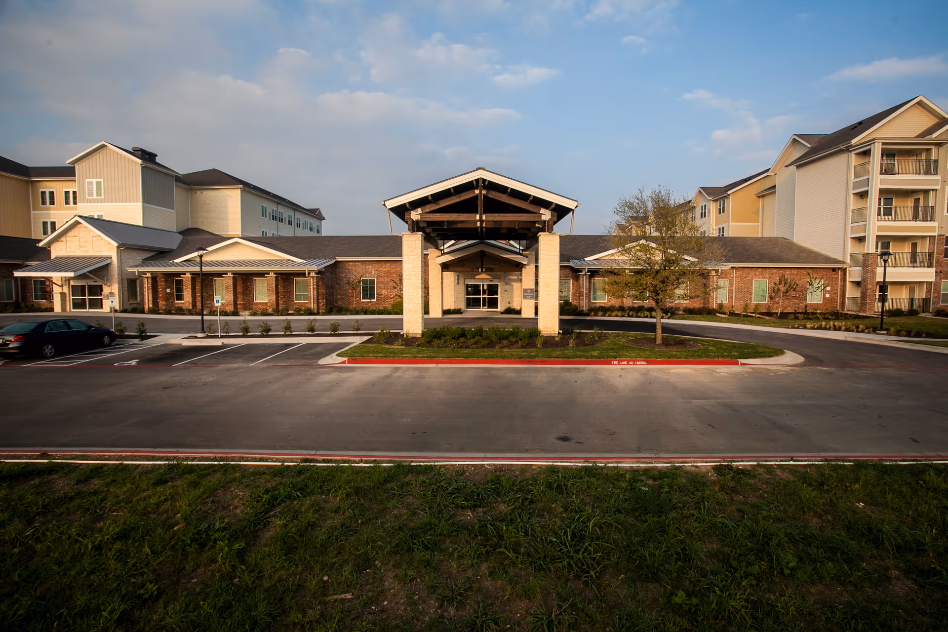Front exterior view of The Delaney at Lake Waco facility showing a large entrance with a covered driveway, surrounded by a parking lot and multi-story buildings under a partly cloudy sky.