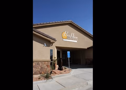 Entrance and facade of the Beehive Homes building with its sign above the doorway, a handicap parking sign, and desert landscaping under a clear blue sky.