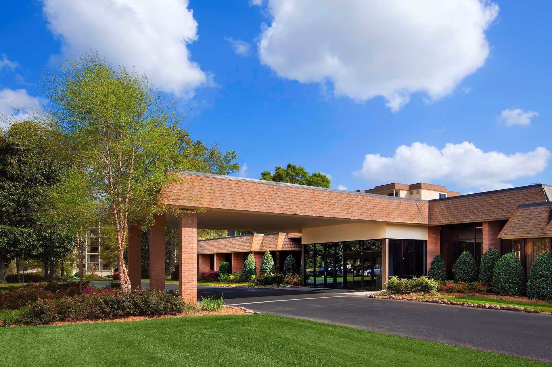 Exterior view of a senior living facility named Azalea Trace, showing a covered entrance with brick pillars, well-maintained landscaping with green grass, bushes, and trees under a blue sky with some clouds.
