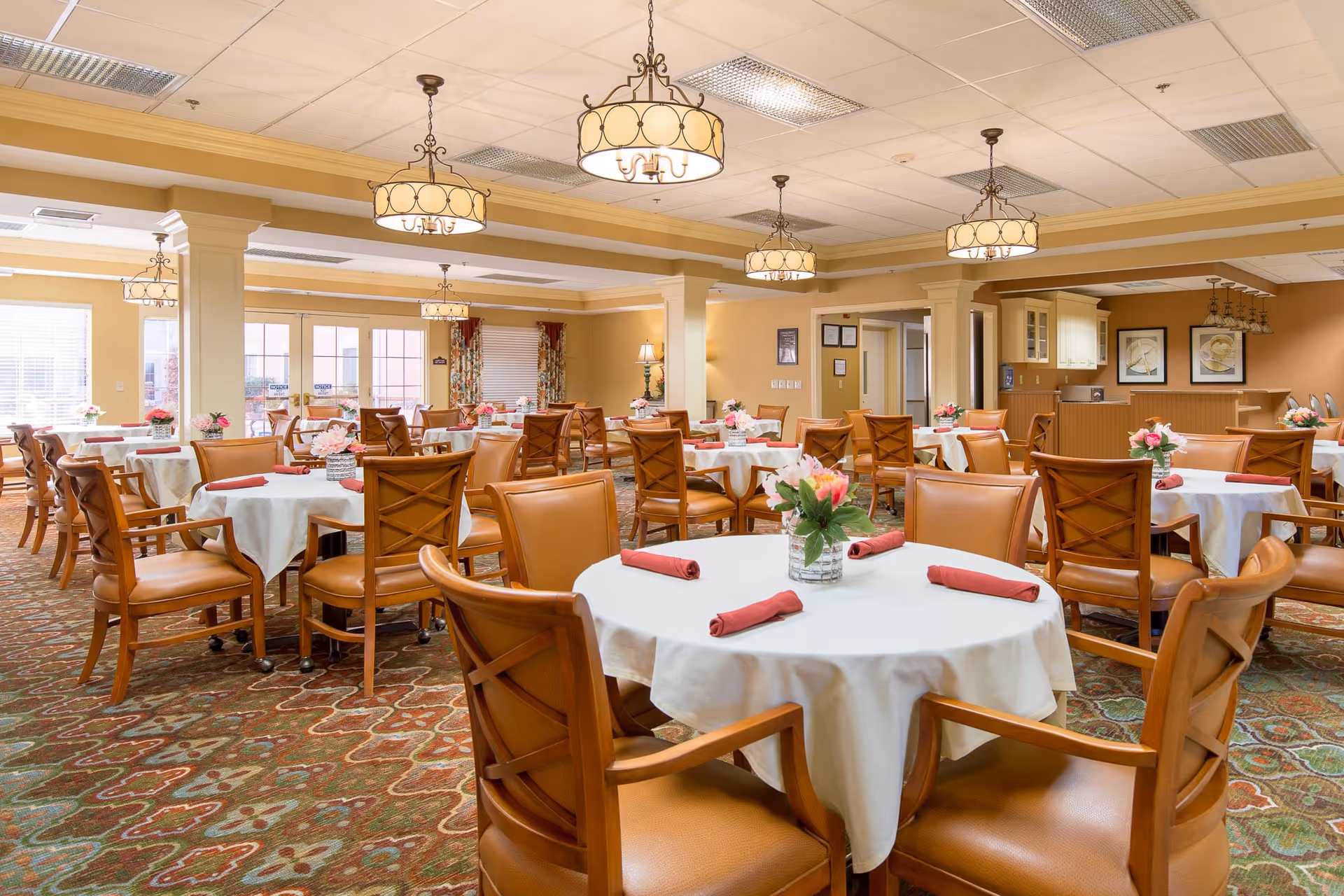 A spacious dining room with multiple round tables covered with white tablecloths, each set with rolled red napkins and a vase of pink flowers. The room features warm beige walls, patterned carpet, wooden chairs with leather seats, and several hanging light fixtures. Large windows and glass doors allow natural light to enter the room.