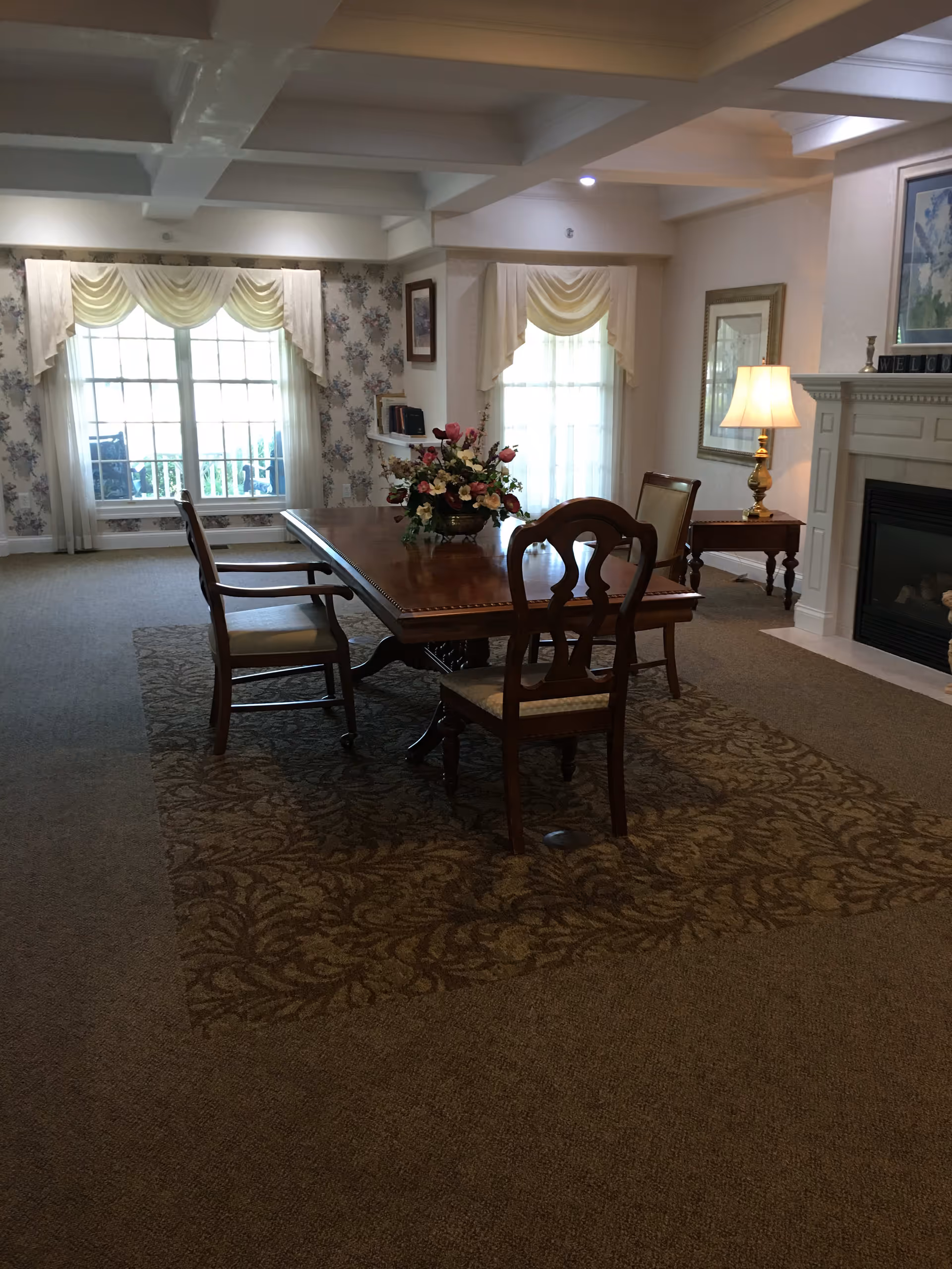 A cozy interior room with a wooden dining table and four chairs placed on a patterned carpet. The room features large windows with sheer curtains and valances, floral wallpaper, a lit table lamp on a side table, and a white fireplace with decorative items on the mantel.