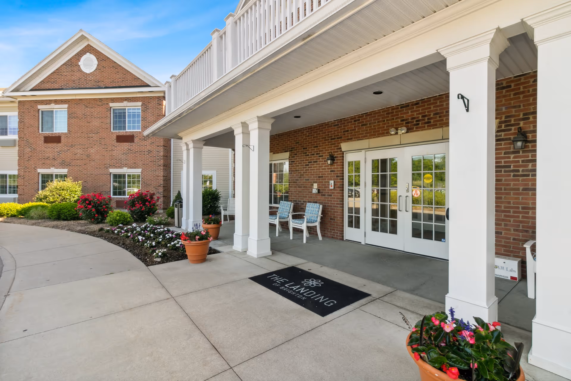 Entrance to The Landing of Brighton facility showing a covered porch with white columns, two white chairs with blue cushions, potted plants with flowers, a brick exterior wall, glass double doors, and a welcome mat with the facility's name.