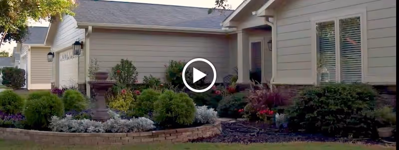 Exterior view of a single-story residential building with beige siding and a gray roof, surrounded by well-maintained landscaping including bushes, flowers, and a small raised garden bed with a stone border.