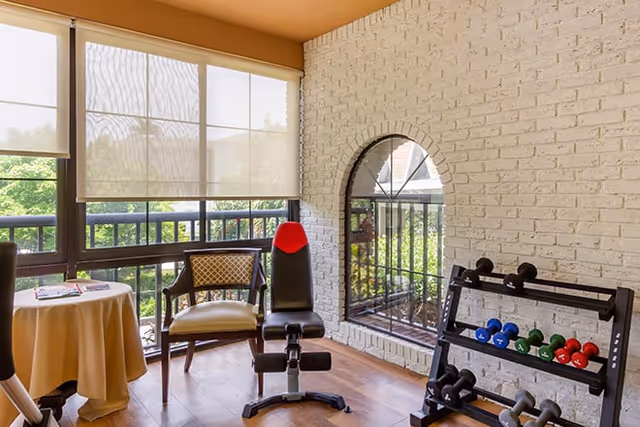 A bright room with large windows covered by white roller shades, a small round table with a beige tablecloth and magazines, a wooden chair with a cushion, a black and red exercise bench, and a rack holding various colored dumbbells. The room has a white brick wall with an arched window opening to an outdoor view.