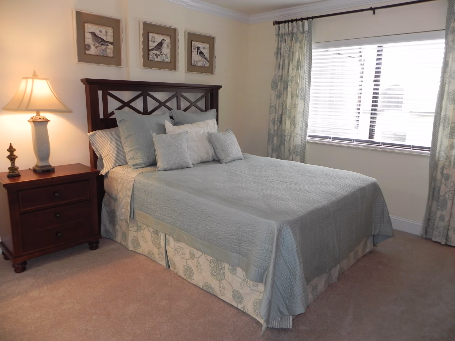 Neatly made bed with light blue bedding, wooden headboard, nightstand with a lamp, framed bird pictures on the wall, and a window with blinds.