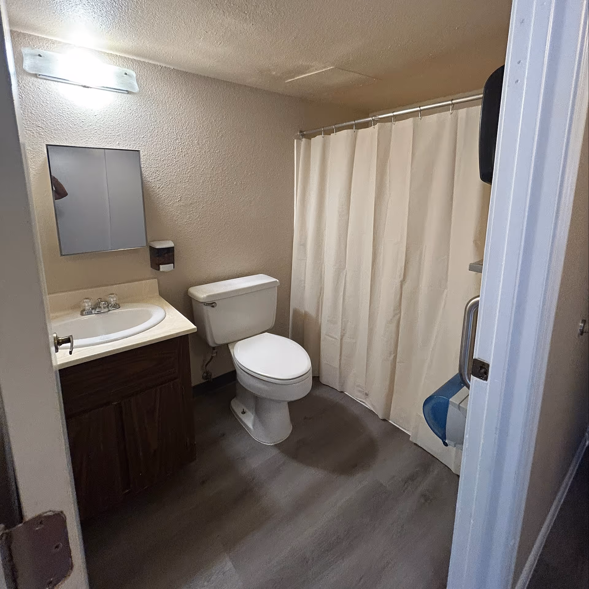 A small bathroom with a white toilet, a sink with a wooden cabinet, a mirror above the sink, a wall-mounted soap dispenser, and a shower with a white curtain. The floor has a grayish tone and the walls are beige.