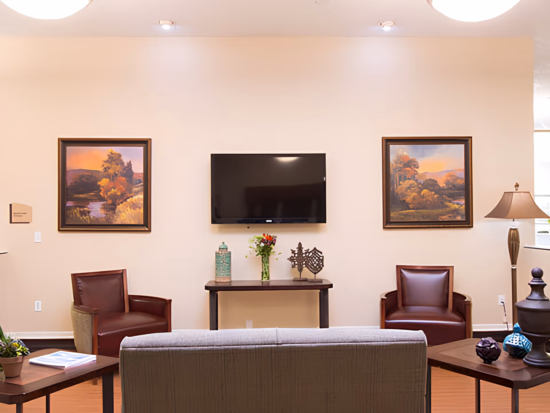Common living room with a wall-mounted TV, two framed landscape paintings, seating, and side tables with decorative items.
