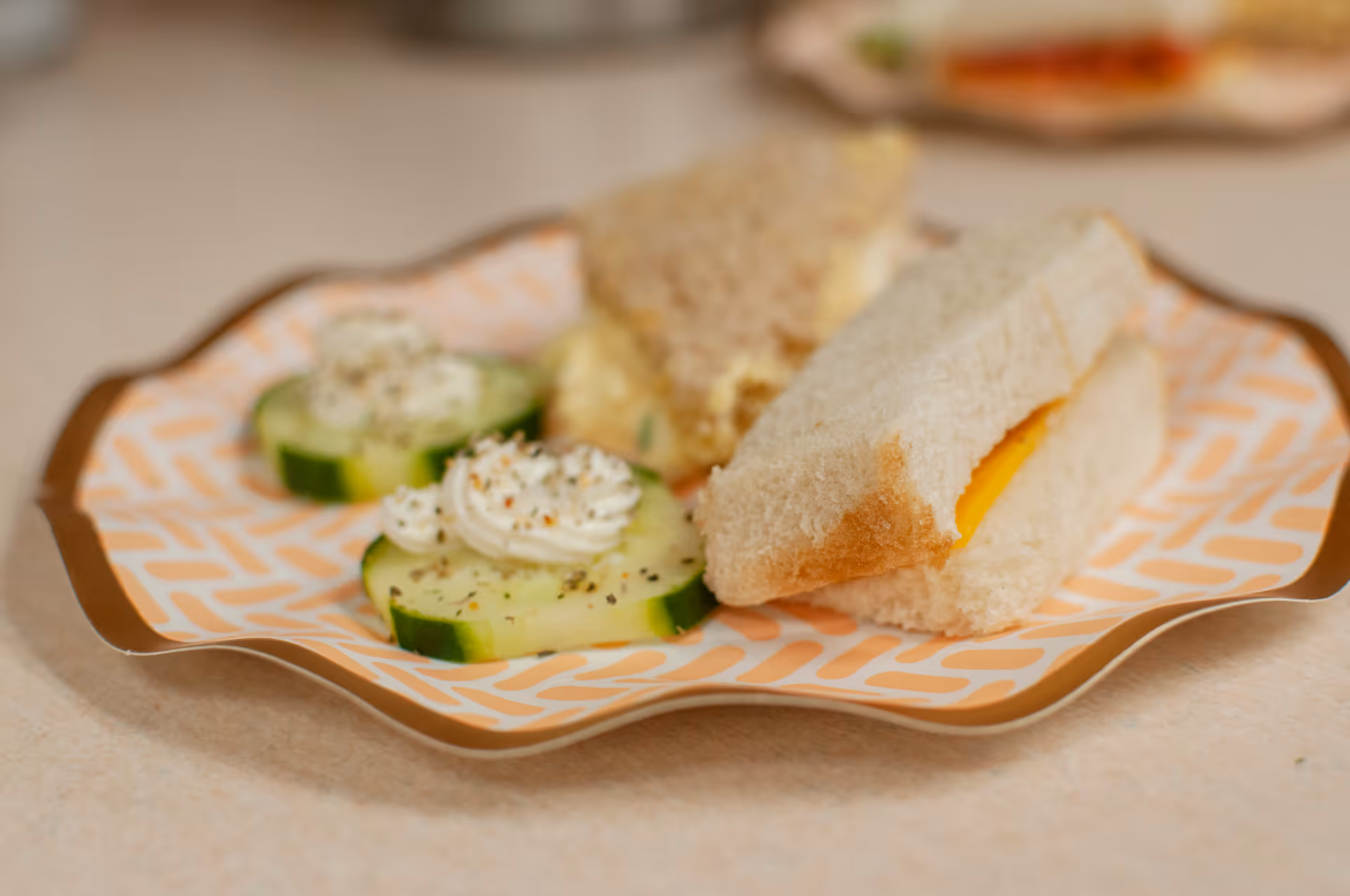 A decorative plate with finger sandwiches and cucumber slices topped with a dollop of cream cheese and sprinkled with black pepper.