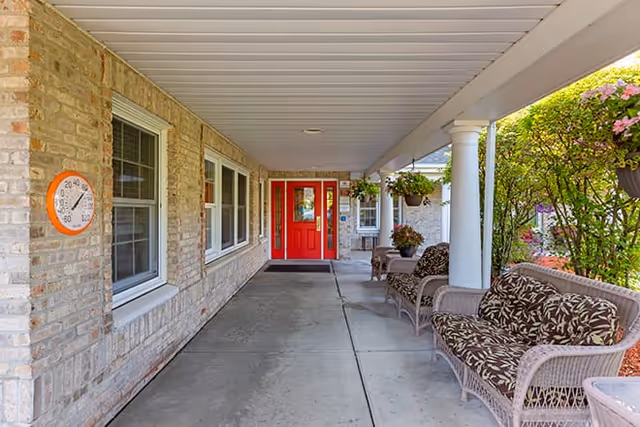 Covered porch leading to a red double-door entrance with wicker chairs and hanging plants.