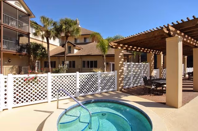 Outdoor area of Vitality Living Hudson featuring a small round pool with metal handrails, a white lattice fence, palm trees, and a pergola with outdoor seating. The building exterior with balconies and windows is visible in the background under a clear blue sky.