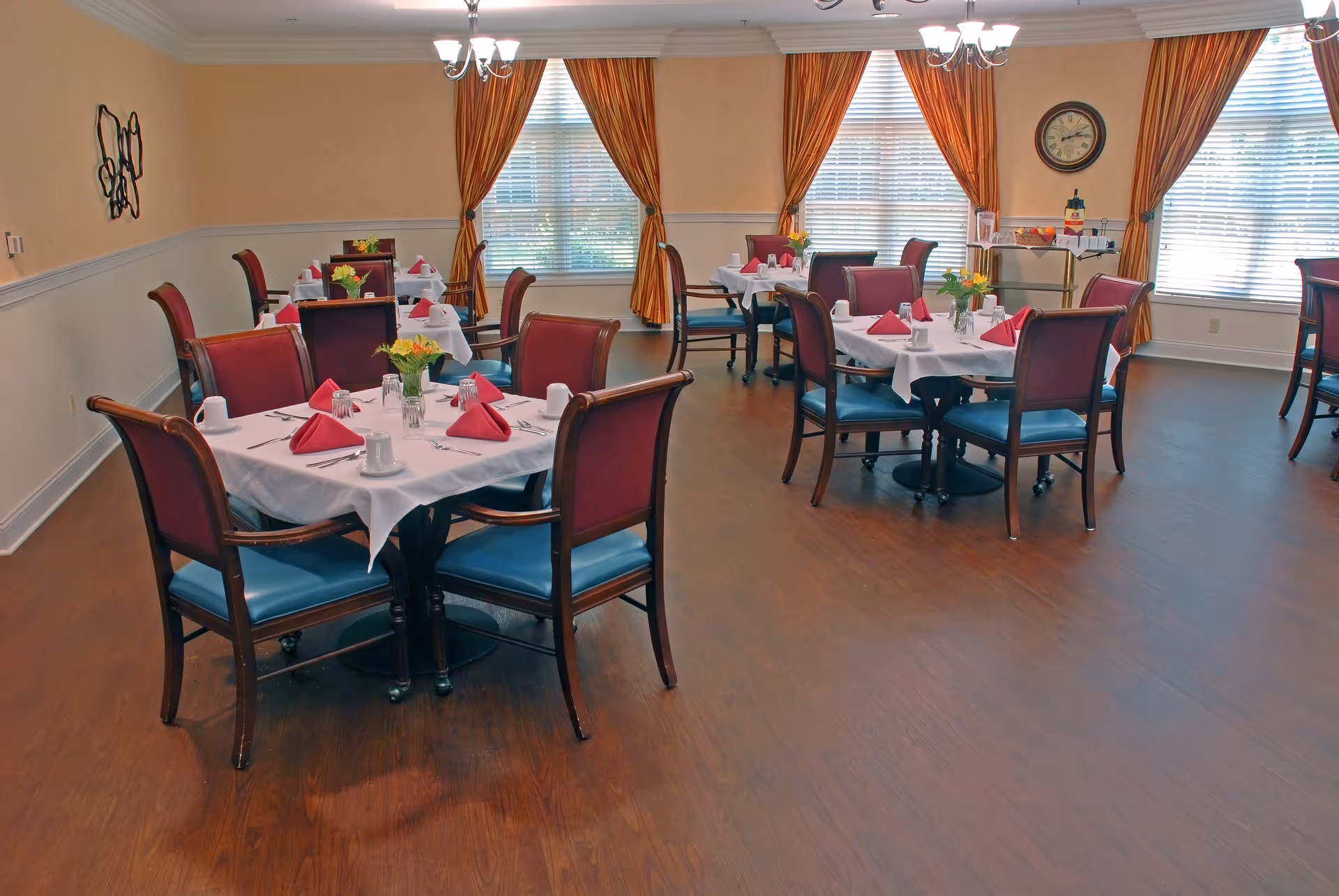 A dining room with multiple tables set for meals, each table covered with a white tablecloth, red folded napkins, cups, and small flower arrangements. The room has wooden flooring, large windows with orange curtains, and a clock on the wall.