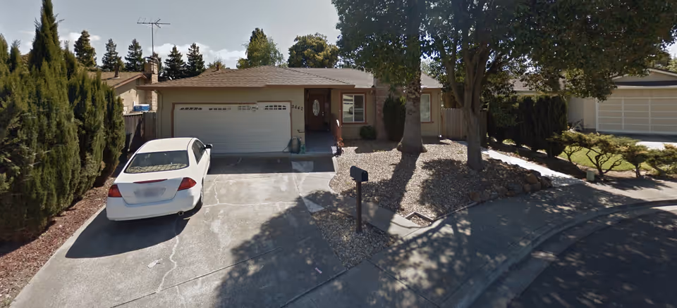 Front exterior view of a single-story house with a two-car garage, a white car parked in the driveway, and a landscaped front yard with trees and shrubs.