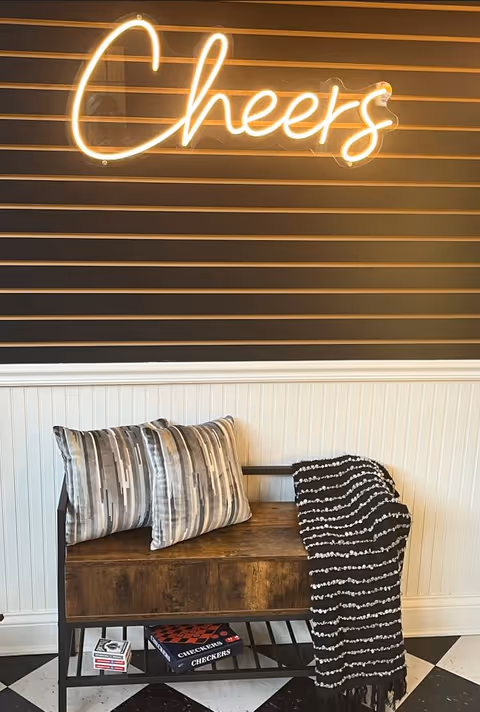 A cozy corner with a wooden bench featuring two patterned pillows and a black and white striped throw blanket. Above the bench, a neon sign glows with the word 'Cheers' mounted on a dark wall with horizontal slats. Below the bench, there are board games including Checkers and a deck of cards. The floor has a black and white checkered pattern.