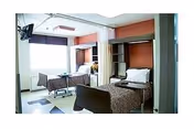 A bright nursing facility room with two hospital beds separated by a curtain. Each bed has pillows and blankets, and there is a window letting in natural light. A television is mounted on the wall near the ceiling.