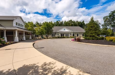 View of the driveway and entrance area of a senior living facility with two buildings surrounded by trees and landscaped gardens under a partly cloudy sky.