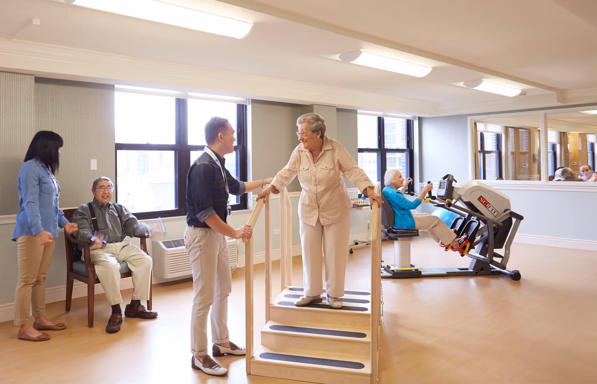 A senior woman uses a wooden step exercise device with the assistance of a caregiver in a bright room. Another elderly woman is exercising on a seated leg press machine in the background. Two other people, one seated and one standing, are observing and interacting in the room with large windows and light-colored flooring.