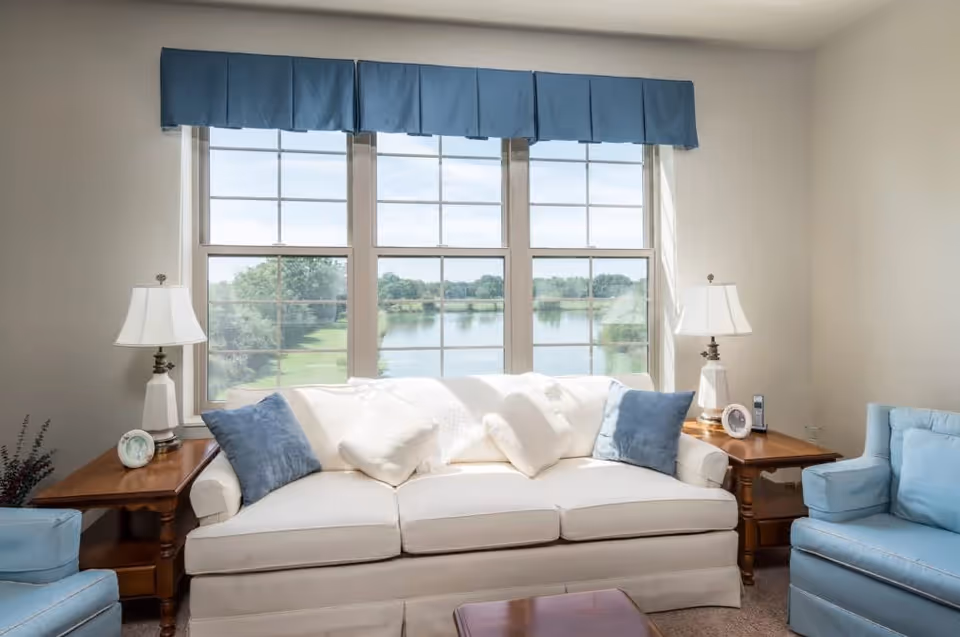 A bright living room with a large window overlooking a scenic view of a lake and trees. The room features a white sofa with blue and white pillows, two wooden side tables each with a white lamp, and two blue armchairs on either side of the sofa.