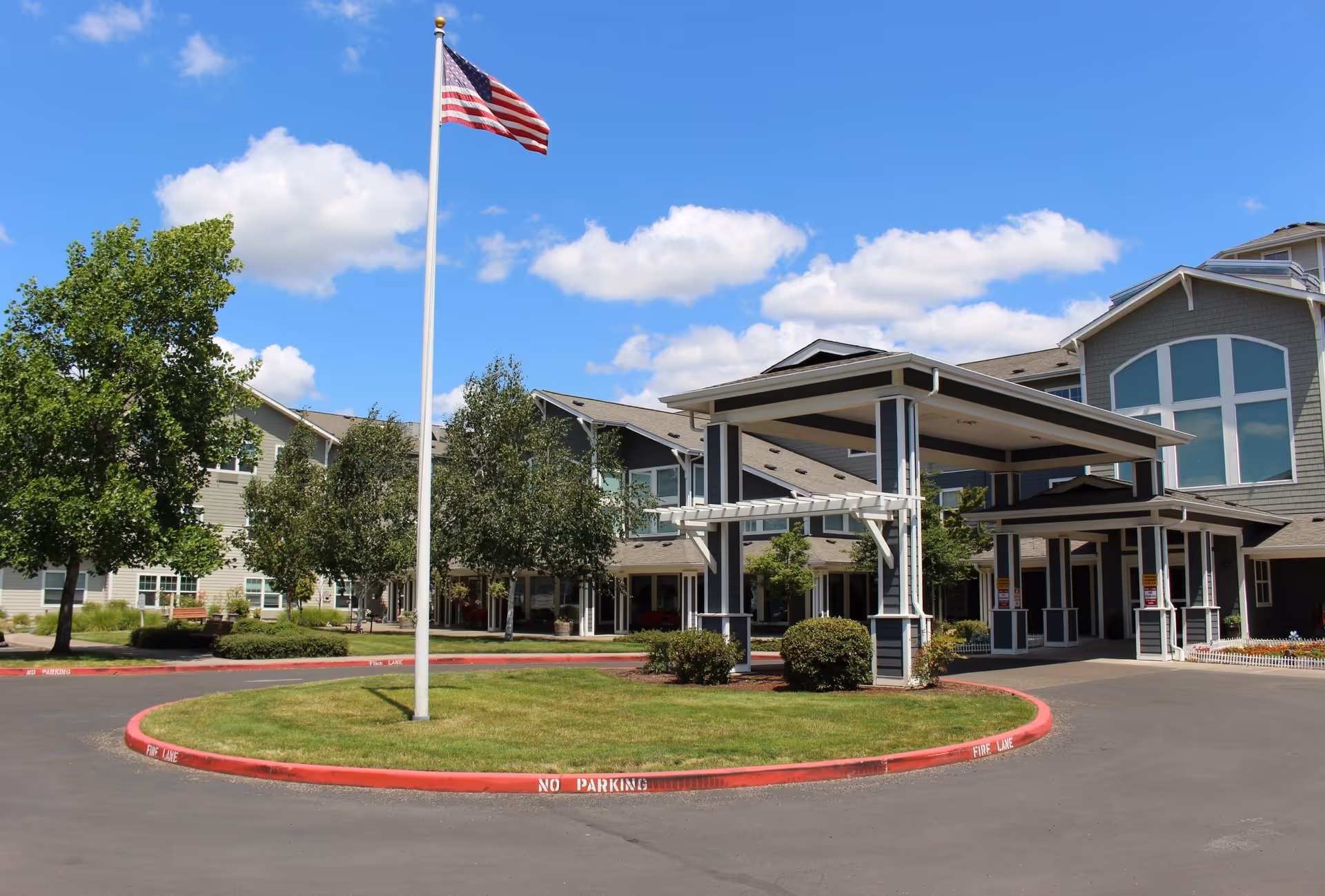 Exterior view of Timber Pointe Senior Living facility showing a large building with multiple windows, a covered entrance with pillars, a circular driveway with a grassy island in the center, and an American flag on a flagpole. The sky is blue with scattered clouds and there are trees and shrubs around the building.
